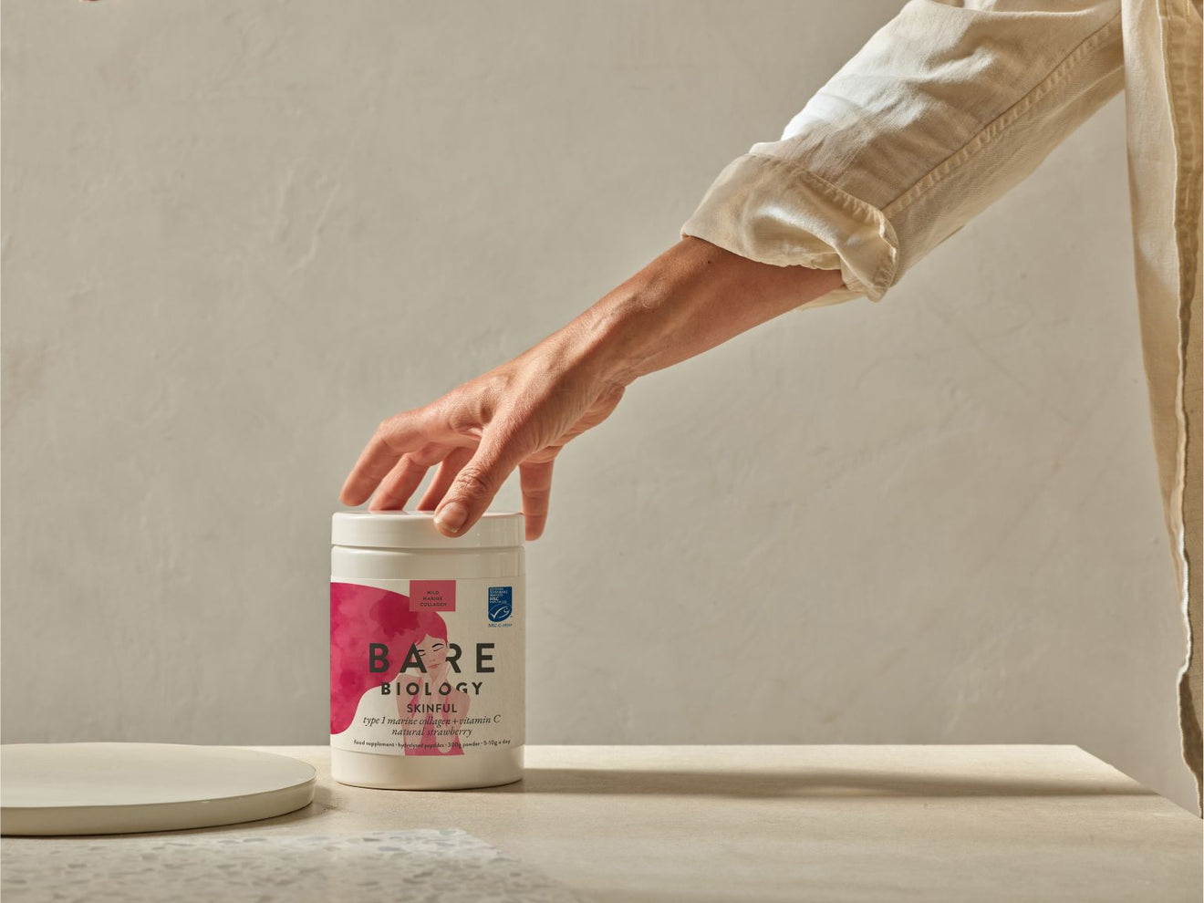 Person holding a container labeled 'Bare Biology Skinful and  Vitamin C Collagen' on a neutral background