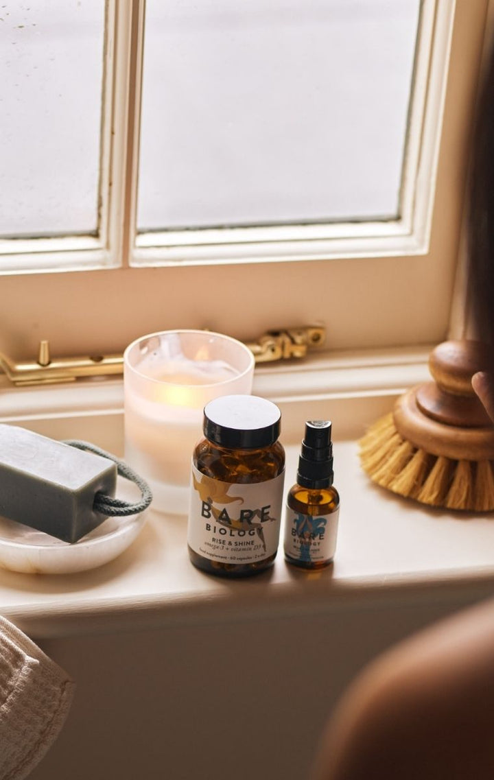 A bathroom or spa setting by a window with a jar labeled 'BARE BIOLOGY' and a small spray bottle beside a lit candle, soap dish, soap bar, and wooden brush.