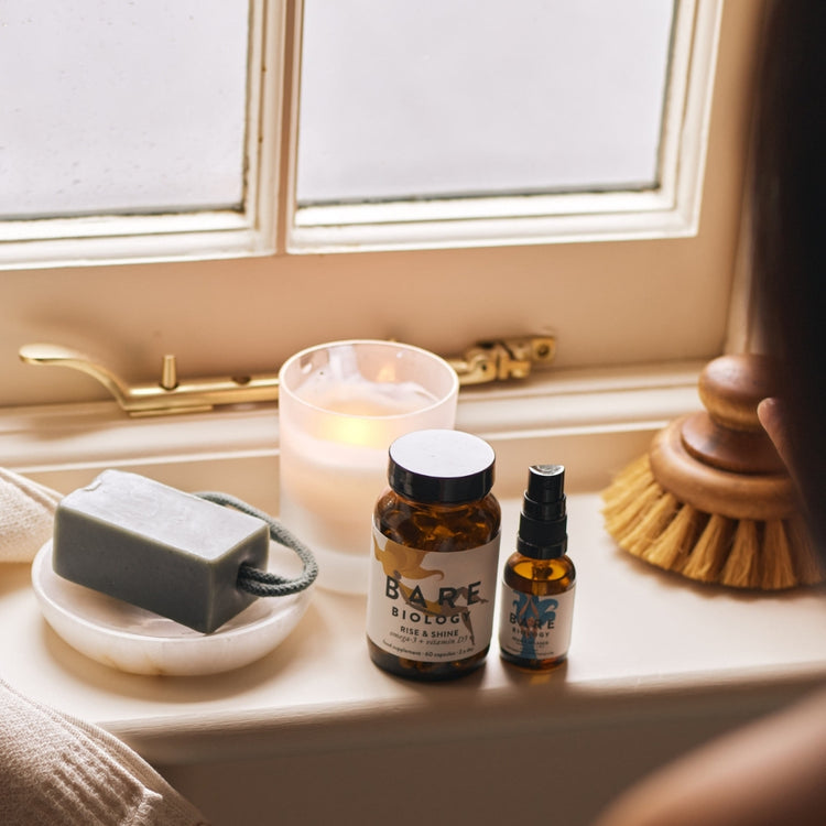 Bath products including a candle, soap, and bottles on a windowsill, with a jar of Bare Biology Rise & Shine omega-3 + vitamin D capsules and vitamin D spray.