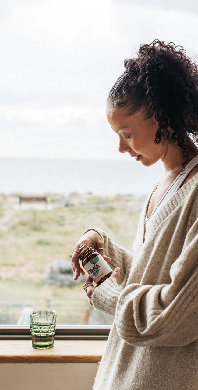 a woman in a cardigan holding mindful omega 3 fish oil capsules next to a window with a view of the sea