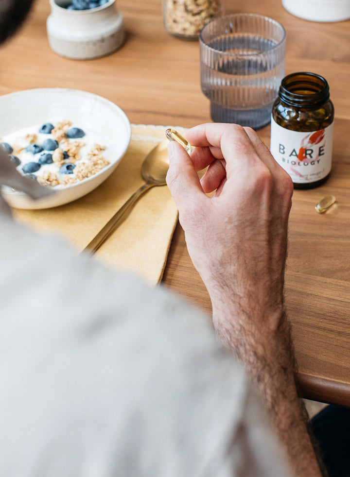 man's hand holding a bare biology omega-3 capsules with a breakfast setting  in the background