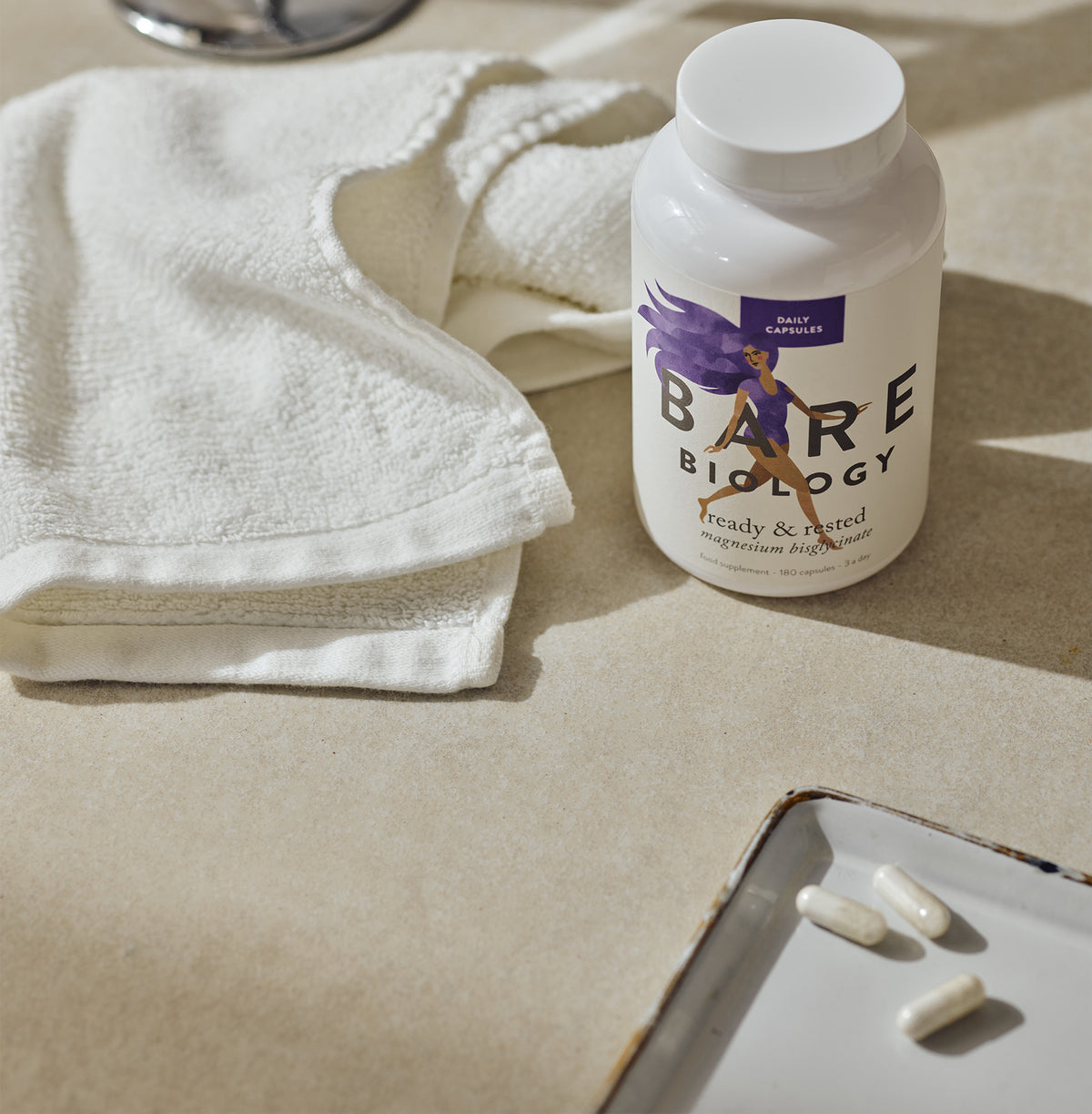 a white jar of bare biology magnesium capsules on a beige stone surface with shadows. There's a white face cloth crumpled beside them and a tray with 3 magnesium capsules in the front corner.
