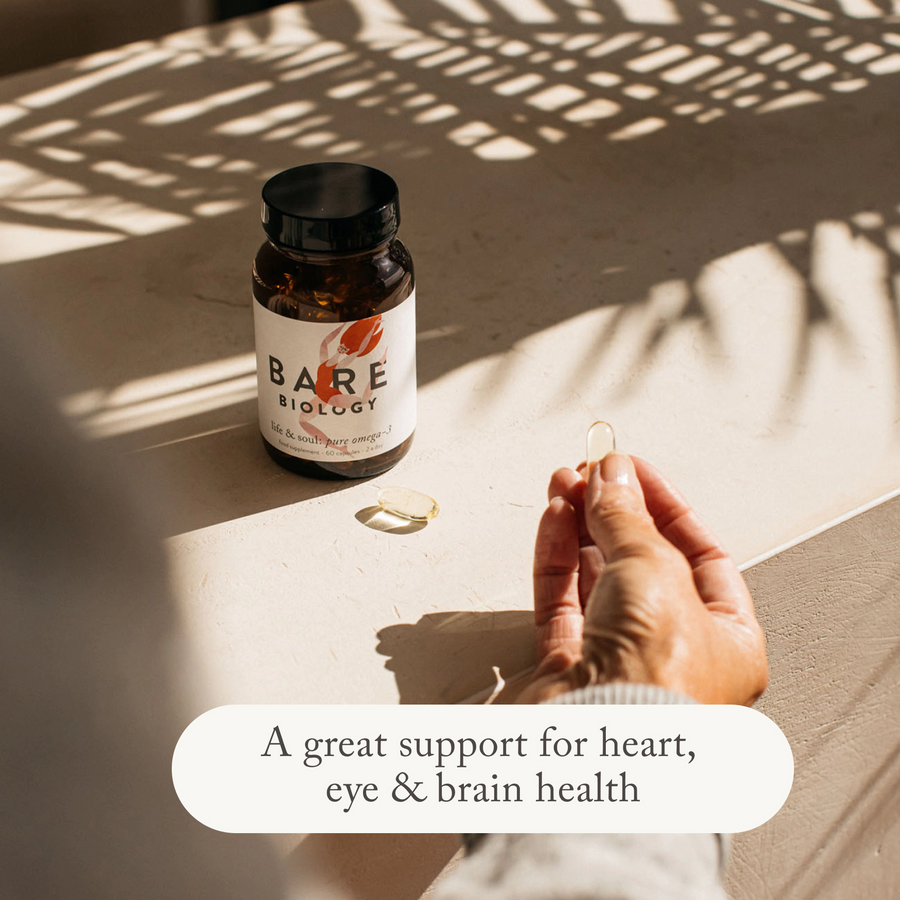 A woman holding a bare biology life & soul daily omega 3 fish oil capsule on a sunny table with shadows of a plant