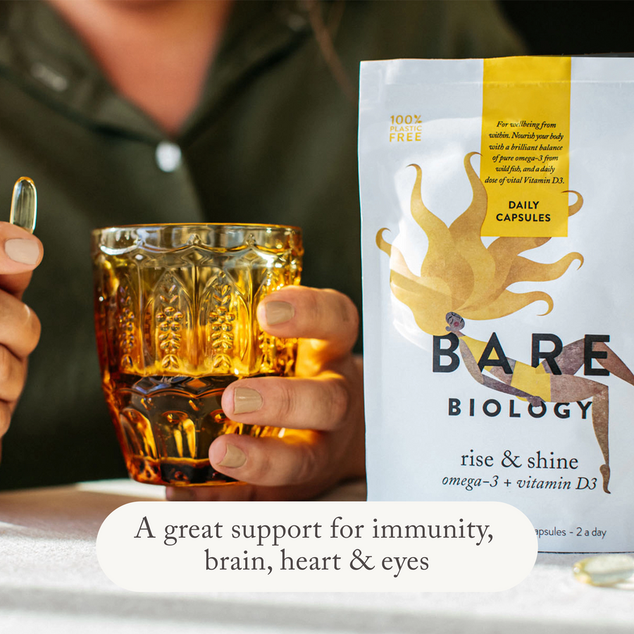a woman holding a bare biology rise & shine vitamin d3 and omega 3 capsule with a glass of water in yellow cup